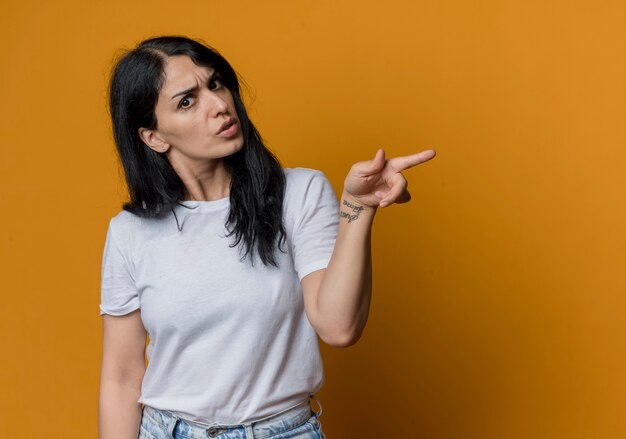 Confused young brunette caucasian girl points at side looking isolated on orange wall