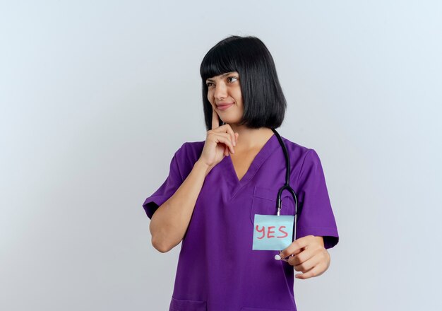 Confused young brunette female doctor in uniform with stethoscope puts hand on chin and holds yes note looking at side 