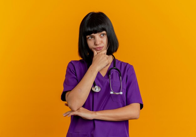 Смущенная молодая брюнетка женщина-врач в униформе со стетоскопом кладет руку на подбородок, глядя в сторону