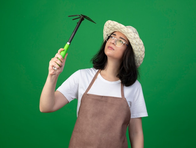 Confused young brunette female gardener in optical glasses and uniform wearing gardening hat holds and looks at hoe rake isolated on green wall