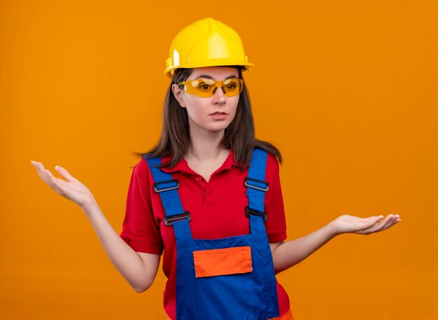 安全メガネで混乱している若いビルダーの女の子は、孤立したオレンジ色の背景に手を上げる