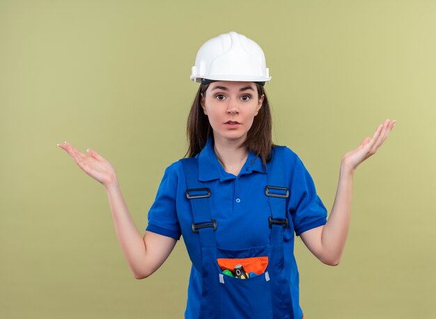 Confused young builder girl with white safety helmet and blue uniform holds hands up and loks at camera on isolated green background