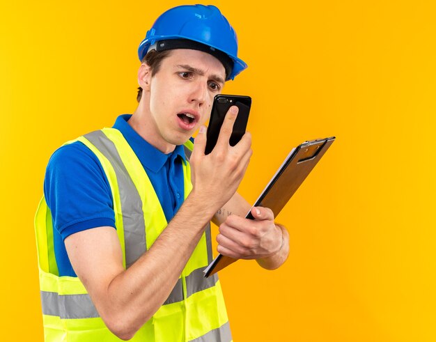 Confused young builder man in uniform holding clipboard and looking at phone in his hand 