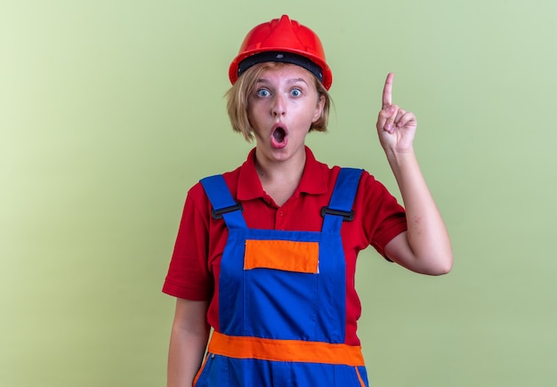 Confuso giovane donna costruttore in uniforme punta verso l'alto isolato sul muro verde oliva