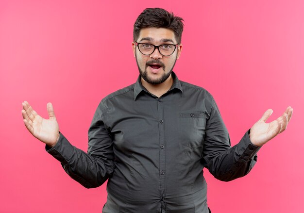Confused young businessman wearing glasses spreads hands isolated on pink wall