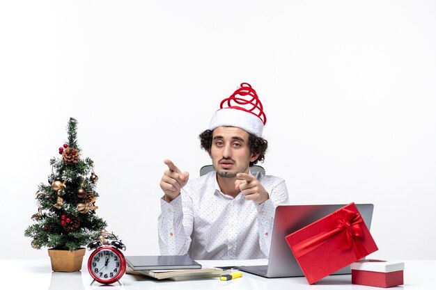 Confused young businessman with funny santa claus hat pointing something in the office on white background