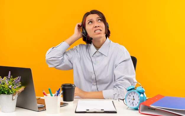 Confused young call center girl wearing headset sitting at desk putting hand on head looking up isolated on orange 