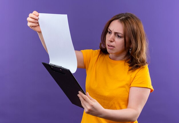 Confused young casual woman holding clipboard and looking at it on isolated purple space