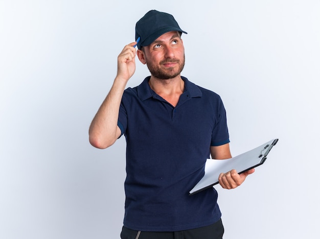 Confused young caucasian delivery man in blue uniform and cap holding clipboard looking up touching head with pen isolated on white wall with copy space