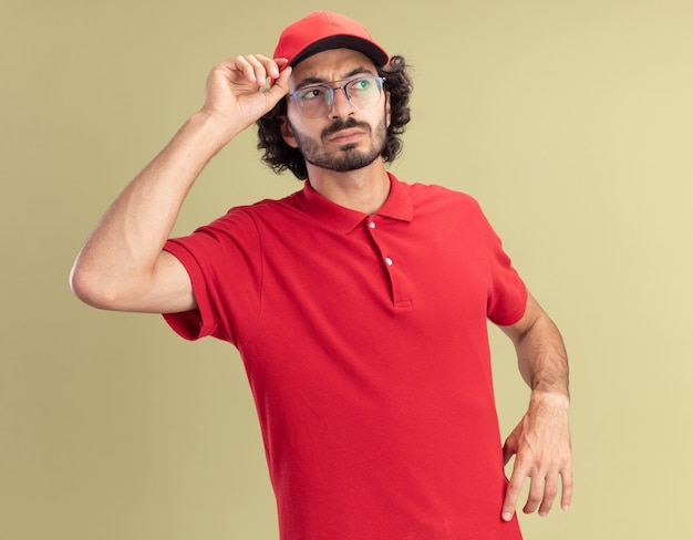 Confuso giovane fattorino caucasico in uniforme rossa e berretto con gli occhiali che afferra il berretto guardando a lato