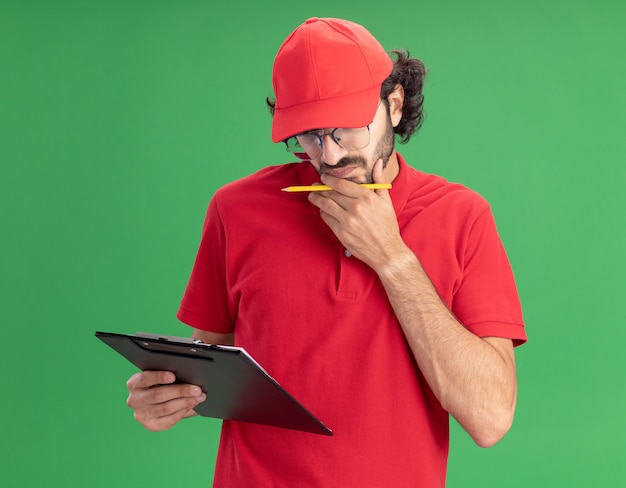 Foto gratuita confuso giovane fattorino caucasico in uniforme rossa e berretto con gli occhiali che tengono appunti e matita mettendo la mano sul mento guardando gli appunti isolati sul muro verde