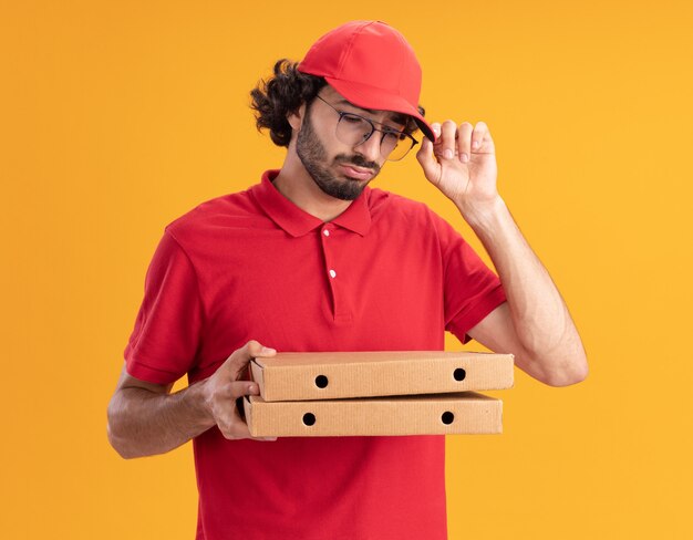 Confused young caucasian delivery man in red uniform and cap wearing glasses holding and looking at pizza packages grabbing cap 