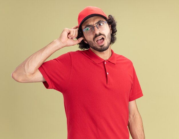 Confused young caucasian delivery man in red uniform and cap wearing glasses looking at side touching head isolated on olive green wall