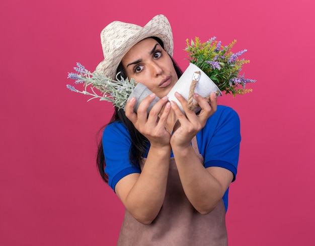 Free photo confused young caucasian gardener girl wearing uniform and hat looking at side holding flowerpots isolated on pink wall