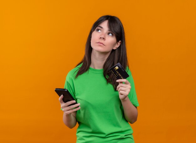 Смущенная молодая кавказская девушка в зеленой рубашке держит телефон и кредитную карту на изолированном оранжевом фоне