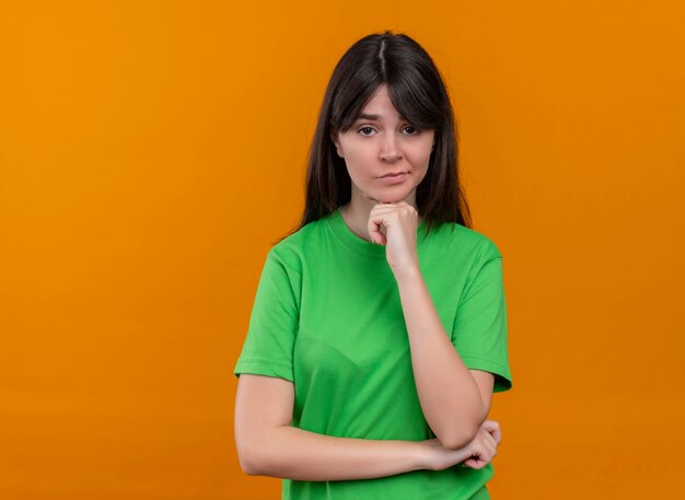 緑のシャツを着た混乱した若い白人の女の子は、あごに手を置き、コピースペースで孤立したオレンジ色の背景にカメラを見る