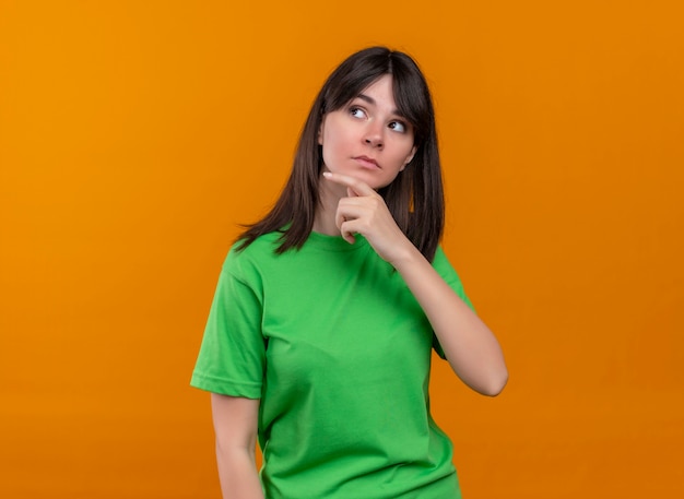 緑のシャツの混乱した若い白人の女の子は、あごに手を置き、孤立したオレンジ色の背景で横に見えます