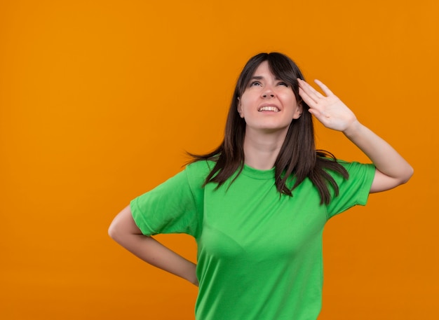 Confused young caucasian girl in green shirt puts hand on head on isolated orange background with copy space