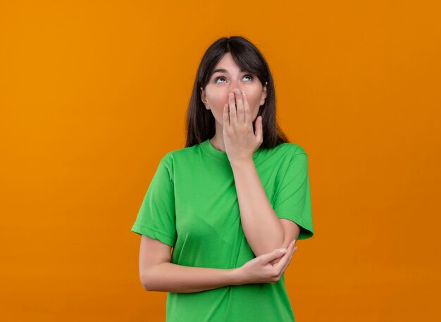 Confused young caucasian girl in green shirt puts hand on mouth and looks up on isolated orange background