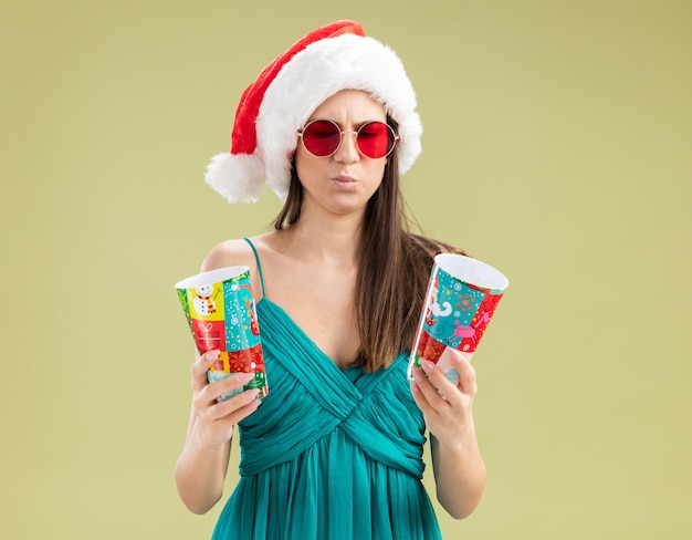 Confused young caucasian girl in sun glasses with santa hat holding and looking at paper cups 