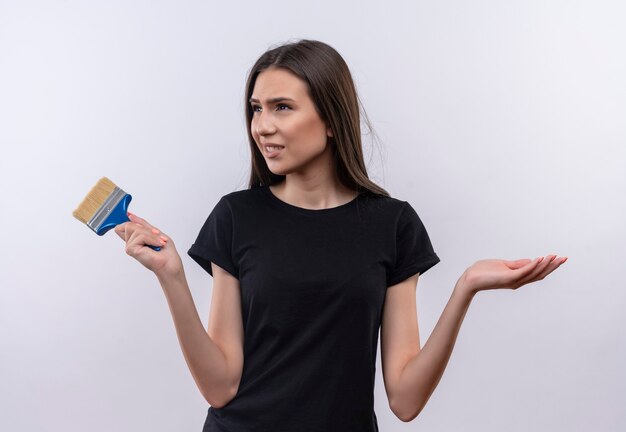 confused young caucasian girl wearing black t-shirt holding paint brush showing what gesture on isolated white wall