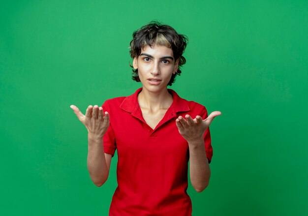 Confused young caucasian girl with pixie haircut showing empty hands isolated on green background with copy space