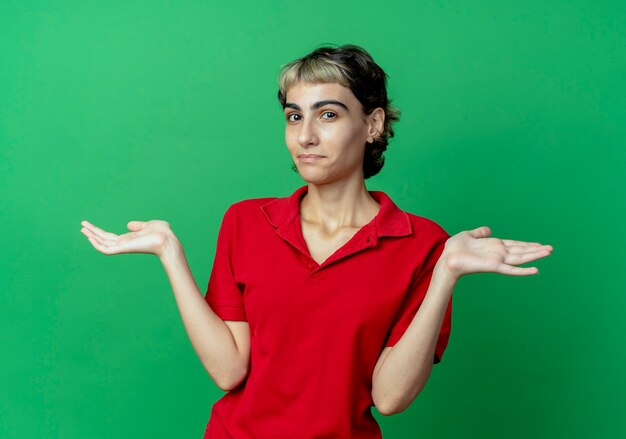 緑の背景に分離された空の手を示すピクシーヘアカットと混乱した若い白人の女の子