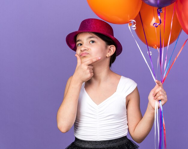 confused young caucasian girl with violet party hat holding helium balloons and putting hand on chin looking up isolated on purple wall with copy space