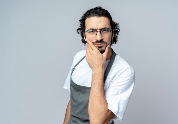 Free photo confused young caucasian male barber wearing glasses and wavy hair band in uniform putting hand on chin isolated on white background with copy space