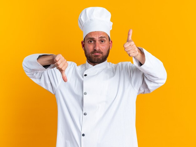 Confused young caucasian male cook in chef uniform and cap looking at camera showing thumbs up and down isolated on orange wall