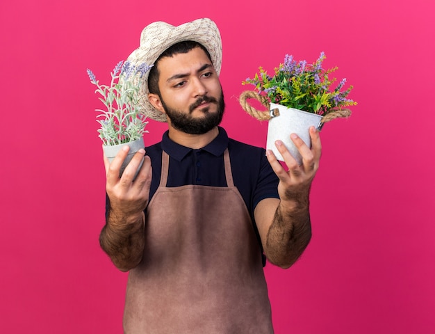 confused young caucasian male gardener wearing gardening hat holding flowerpots isolated on pink wall with copy space