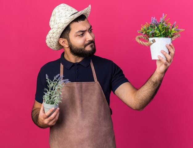 смущенный молодой кавказский садовник в садовой шляпе, держащий и смотрящий на цветочные горшки, изолированные на розовой стене с копией пространства