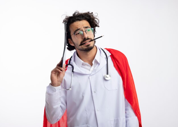 Confused young caucasian superhero man in optical glasses wearing doctor uniform with red cloak and with stethoscope around neck holds clipboard and pencil with teeth isolated on white wall