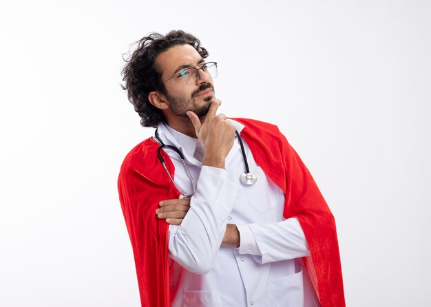 Confused young caucasian superhero man in optical glasses wearing doctor uniform with red cloak and with stethoscope around neck puts hand on chin looking at side with copy space