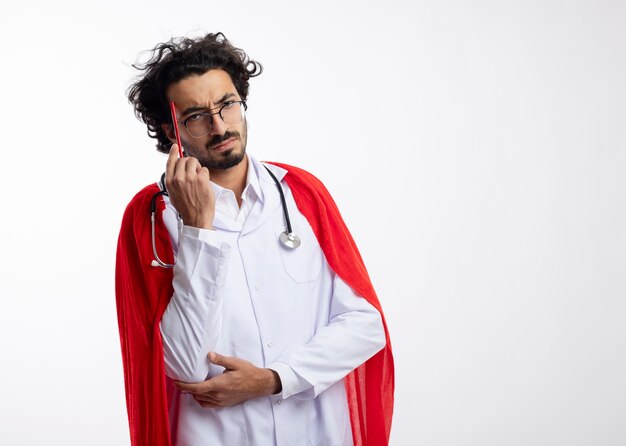 Confused young caucasian superhero man in optical glasses wearing doctor uniform with red cloak and with stethoscope around neck puts pencil on temple 