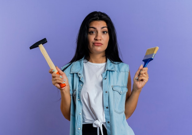 Confused young caucasian woman holds hammer and paint brush isolated on purple background with copy space
