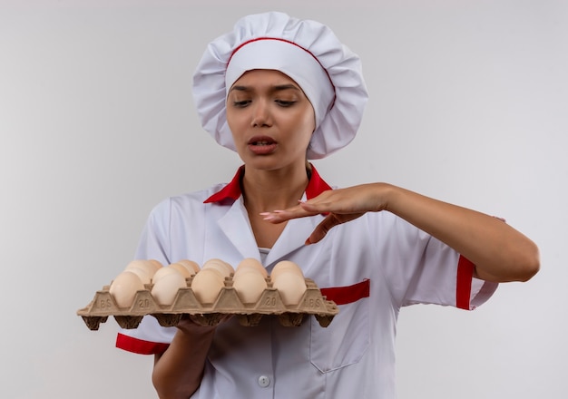 孤立した白い壁に卵のバッチを保持しているシェフの制服を着ている混乱した若い料理人の女性
