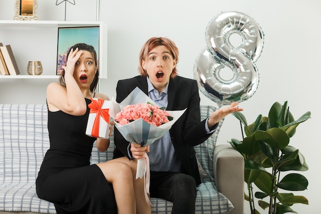 Смущенная молодая пара в счастливый женский день держит подарок с букетом, сидя на диване в гостиной