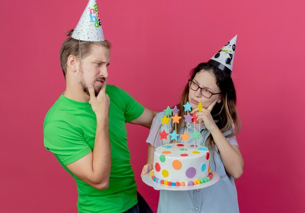 パーティーハットをかぶって混乱している若いカップルはお互いを見て、誕生日の女の子はピンクの壁に隔離されたケーキを保持します