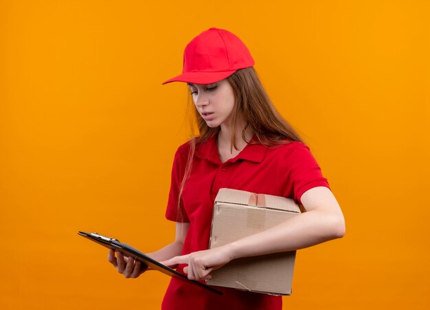Смущенная молодая доставщица в красной форме держит коробку и кладет палец в буфер обмена и смотрит на него в изолированном оранжевом пространстве