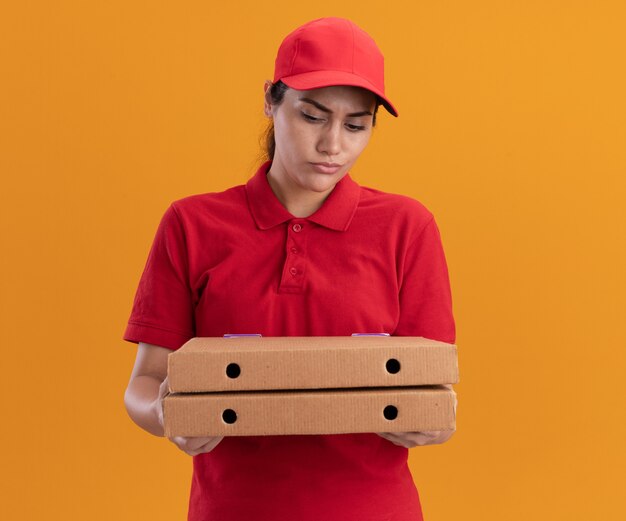 Confused young delivery girl wearing uniform and cap holding and looking at pizza boxes isolated on orange wall