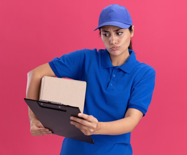 Confused young delivery girl wearing uniform with cap holding box and looking at clipboard in her hand isolated on pink wall