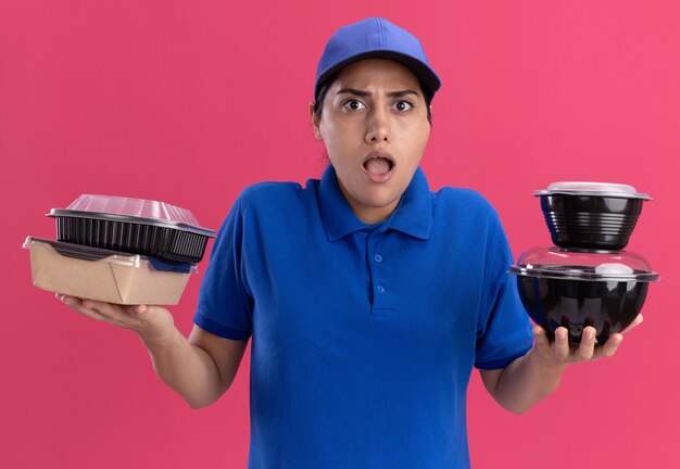 Confused young delivery girl wearing uniform with cap holding food containers isolated on pink wall