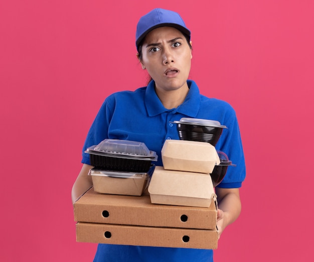 Giovane ragazza confusa delle consegne che indossa l'uniforme con il cappuccio che tiene i contenitori per alimenti sulle scatole per pizza isolate sulla parete rosa