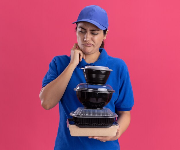 Confused young delivery girl wearing uniform with cap holding and looking at food containers scratching cheek isolated on pink wall