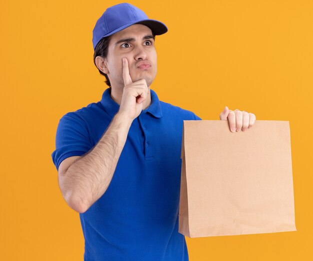 Смущенный молодой курьер в синей форме и кепке держит бумажный пакет, глядя в сторону, положив руку на подбородок, поджимая губы, изолированные на оранжевой стене