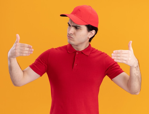 Confused young delivery man in red uniform and cap pretend holding something in front of him looking at his hand isolated on orange wall