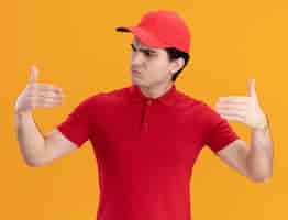 Free photo confused young delivery man in red uniform and cap pretend holding something in front of him looking at his hand isolated on orange wall