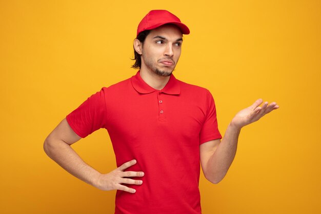 confused young delivery man wearing uniform and cap looking at side showing empty hand while keeping another hand on waist isolated on yellow background