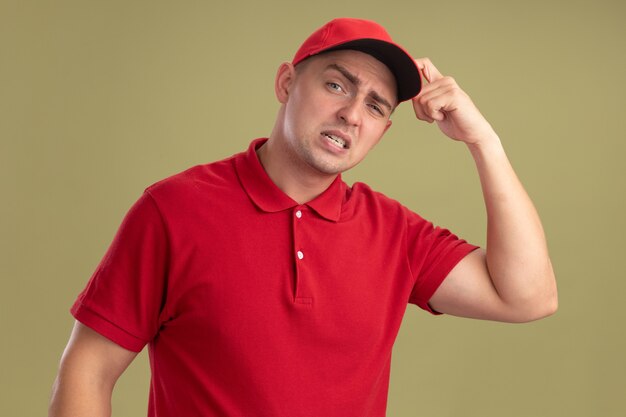 Confused young delivery man wearing uniform and cap scratching head isolated on olive green wall